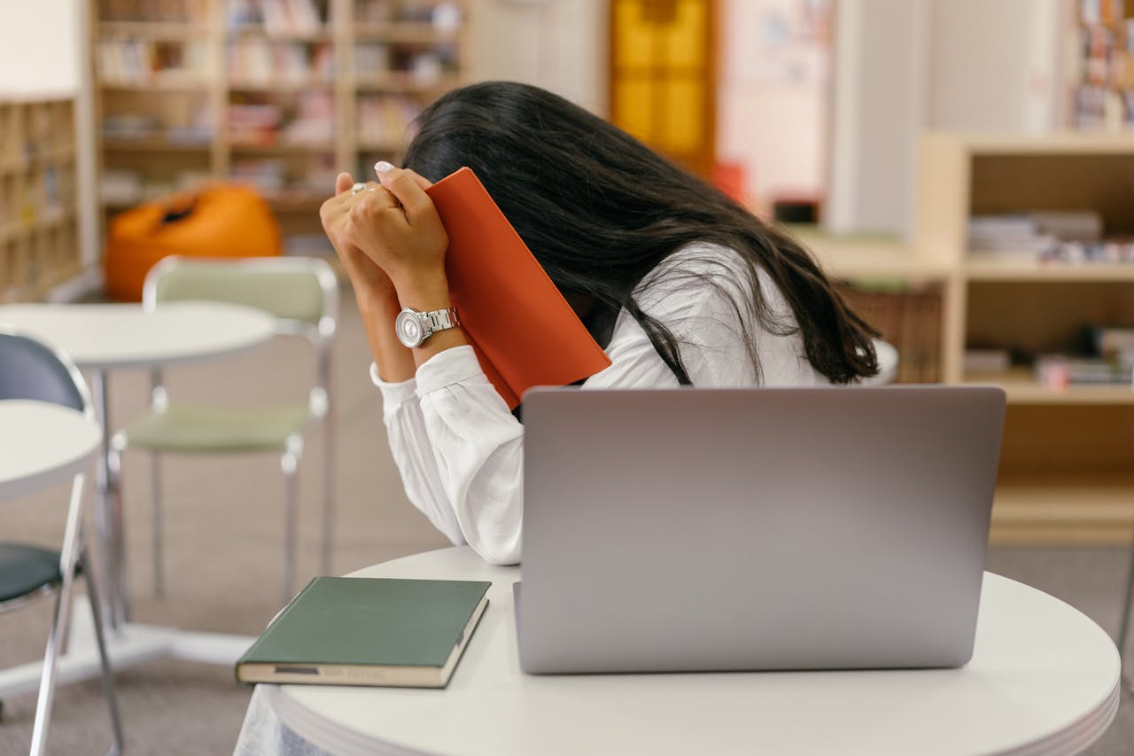 A student hides her face in a book, sitting at a library desk with a laptop, conveying stress.